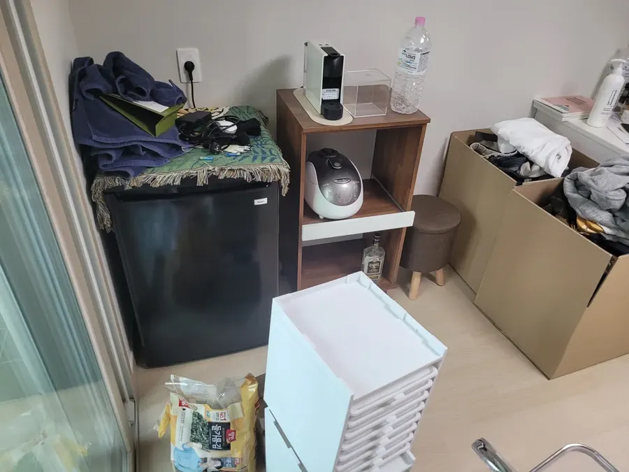 창가에 소형 냉장고와 선반형 수납장, 서랍 수납함, 종이상자가 모여 있는 원룸